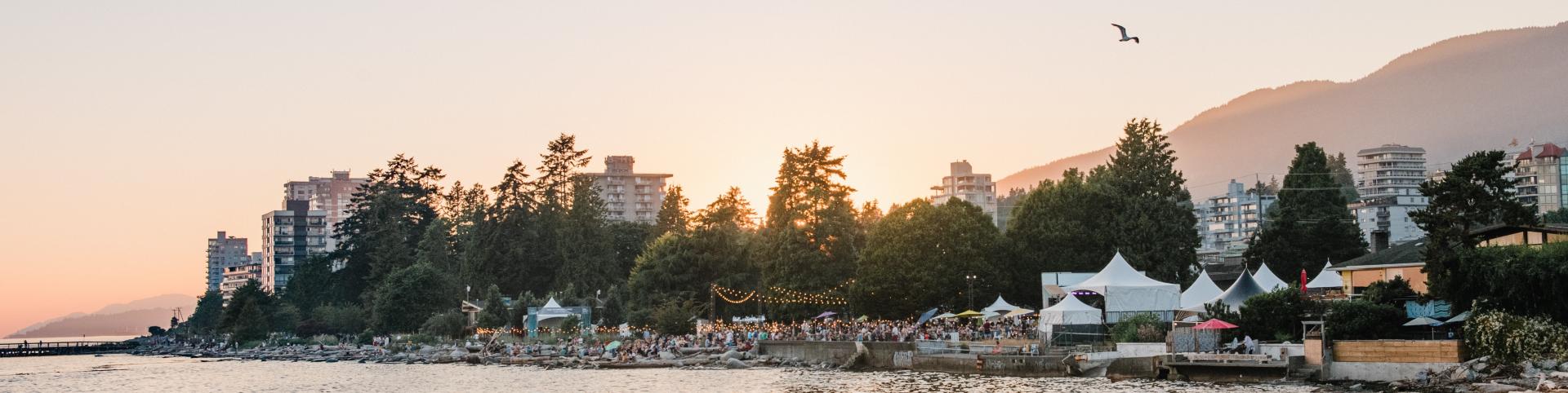 A large crowd gathers on the waterfront, enjoying a summer evening under the setting sun.  The scene is framed by a row of apartment buildings and lush green trees.  The sun casts a warm glow over the water, creating a sense of peace and tranquility.  A boardwalk extends into the water, and several white tents are set up on the shore, suggesting a festival or event.  The scene is alive with laughter and conversation, creating a vibrant and festive atmosphere. 
