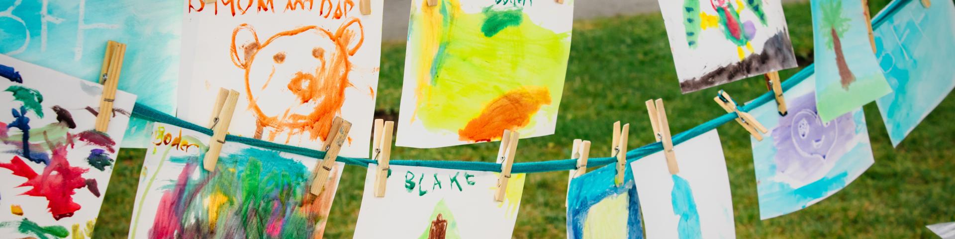 A line of colorful paintings hang on a clothesline strung between two trees. The paintings are all done in watercolor and feature a variety of subjects, including a bear, a tree, and a clown. The paintings are all done by children, and they are a testament to their creativity and imagination. The paintings are displayed outdoors, and the green grass and trees in the background create a beautiful and natural setting. The paintings are a reminder of the simple joys of childhood and the power of art to bring p