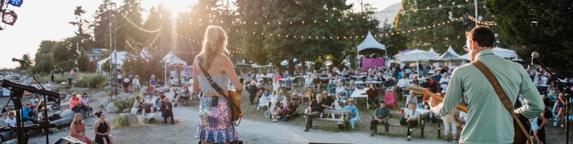 A live music performance takes place outdoors, illuminated by the warm glow of the setting sun. Two musicians, a woman with long blond hair and a man with dark hair, stand on a small stage, facing a crowd of people seated on the ground and benches. The woman is dressed in a sequined skirt and a white tank top, while the man wears a light green shirt and black pants. They both hold guitars, and the man is in the middle of playing his. The stage is equipped with two large speakers and a few microphones. The a