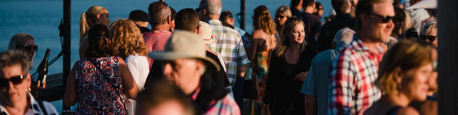 Full shot of a large crowd of people, likely at an outdoor event or gathering by a body of water.   The people are densely packed together, with various ages and styles of clothing.   Many people are wearing casual attire, including shirts, tops, and dresses, and some are visible with hats and sunglasses. 
