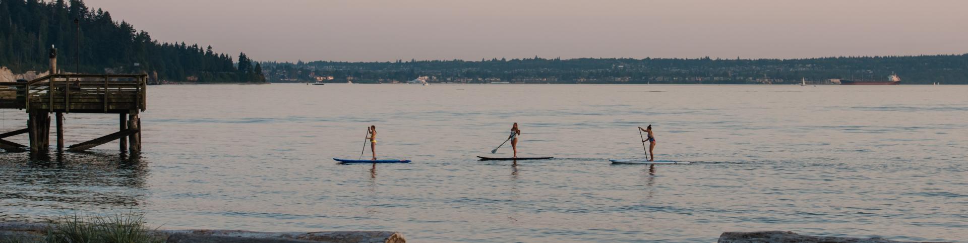 Full outdoor shot of a calm body of water, likely a bay or river.   Three people are stand-up paddleboarding (SUP). They are spaced apart, each with a paddle in their hands. They are positioned in the middle of the water, with a calm, reflective surface around them.
