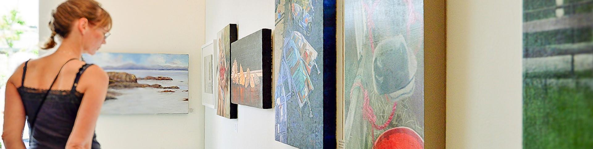 A woman gazes thoughtfully at various paintings displayed on a gallery wall.