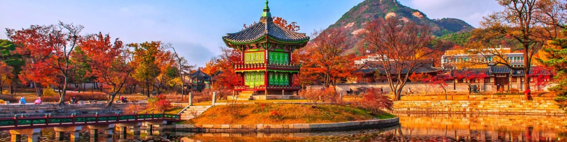 The photo depicts a building, possibly a temple or palace, with a green roof located by a body of water. In the background, there are trees and mountains. This scene is set in Korea.