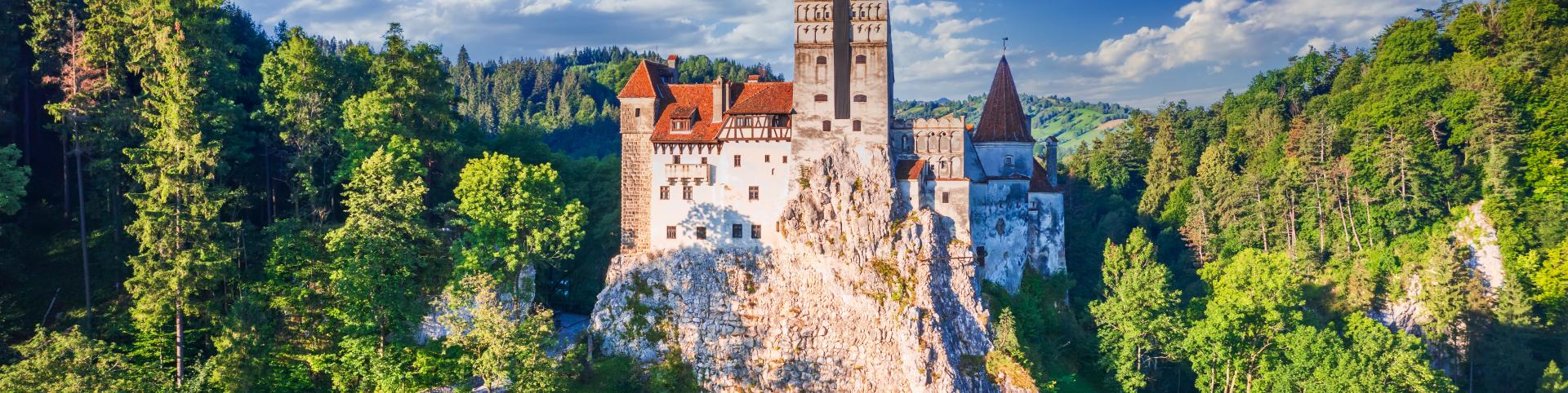 This image shows Bran Castle, a dramatic medieval fortress perched on a rocky hilltop, surrounded by dense green forest under a bright blue sky with scattered clouds. The castle features tall towers with red-tiled roofs and stone walls, giving it a classic Gothic appearance. Bran Castle is famously associated with the Dracula legend and is located in Transylvania, Romania. The lush greenery and mountainous landscape create a picturesque and mysterious setting.