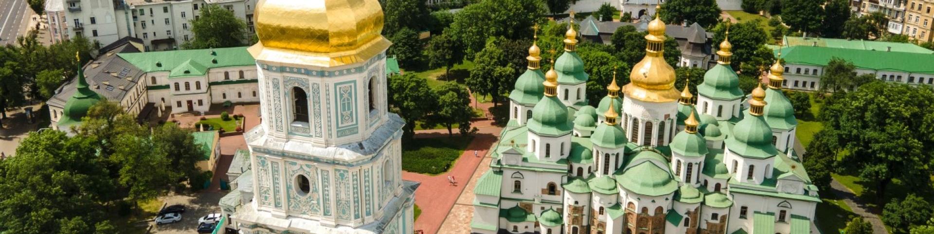 The image is of a large building with a gold-domed roof surrounded by trees. It is a cathedral in Ukraine known as St. Sophia's Cathedral in Kiev, as seen from the height of St. Sophia's Square in the cityscape.