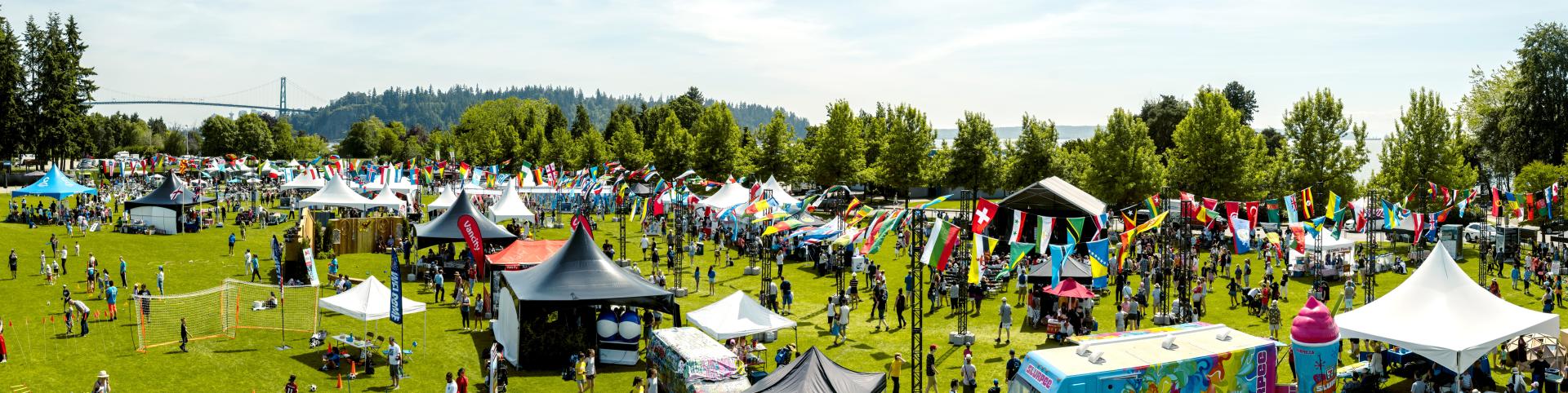 High-angle, full shot of a large outdoor Cultural Fest. A grassy field is filled with numerous tents, canopies, and booths of various colors and sizes. Many people are scattered throughout the field, engaged in activities, and mingling. There are food trucks and various inflatables. 