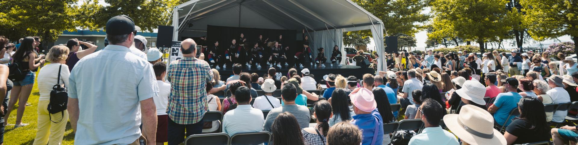 A large crowd of people seated in chairs, enjoying an outdoor dance stage under a clear sky.