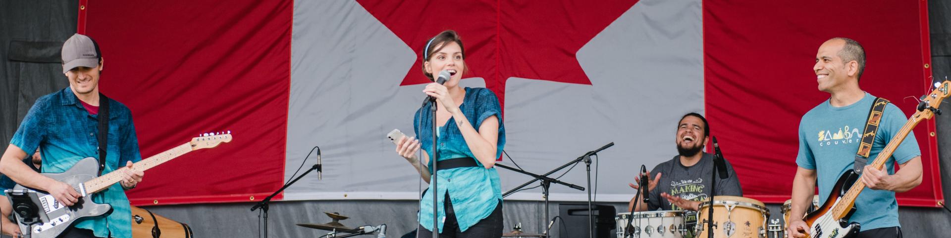 A diverse group of musicians performs on stage, featuring a female singer, male guitarist, bass player, and drummer, with a Canadian flag.