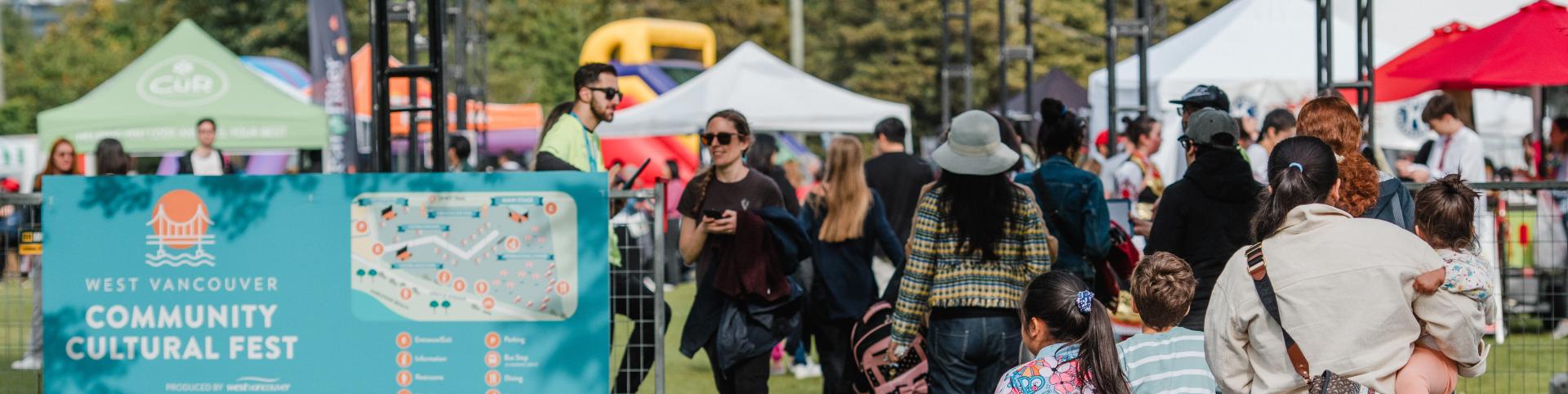  People stroll through a festival entrance adorned with a sign reading "West Vancouver Community Cultural Fest" during a vibrant day.