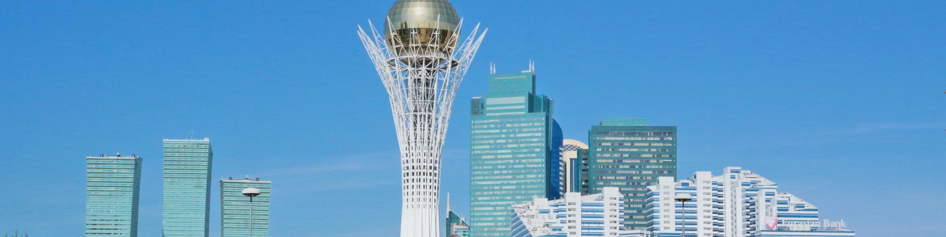 The image depicts a modern cityscape under a clear, bright blue sky. In the foreground, a wide, shallow fountain with multiple jets of water spraying upwards dominates the lower part of the frame. The water is a vibrant turquoise color. The area surrounding the fountain is paved with large, reddish-brown tiles interspersed with dark grey strips, forming a patterned walkway.