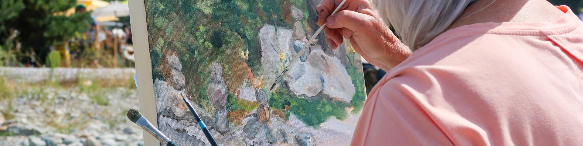 In a close-up, over-the-shoulder shot, an elderly person with white hair is painting outdoors. They are wearing a straw hat that casts a slight shadow over their face, and a salmon-pink t-shirt. The artist holds a thin paintbrush in their right hand, their fingers stained with paint, and is actively working on a canvas set on a dark easel. 