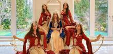 The four surrounding women are dressed in matching elaborate costumes, primarily in rich, deep red velvet with gold accents. Their gowns feature wide, flowing sleeves and ornate gold embroidery on the bodice and waist. Each woman wears a similar headpiece and a sheer, golden veil that flows gracefully around them. Their poses are dynamic and stylized: the two on the left and right are seated with one hand raised near their head, holding the veil, while the other hand is extended outward. 