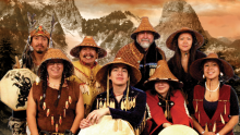 The image is a composite that features a group of nine people, likely Indigenous performers, arranged in two rows against a dramatic backdrop of snow-capped mountains and a fiery, sunset-like sky. The overall color palette is dominated by warm, earthy tones of brown, orange, and gold, with accents of dark green from the pine trees and the blue and red of the masks and some clothing.