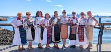 A group of nine women, all of varying ages and ethnicities, are posing for a photo outdoors on a sunny day. They are standing on a boardwalk or pier, with a body of water and a city skyline in the background. The women are dressed in traditional, elaborately embroidered folk costumes. The costumes are predominantly white with intricate red, black, and blue embroidery on the sleeves, necklines, and hems. Many of the women are wearing layered skirts and sashes, also adorned with traditional patterns and brigh