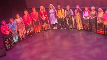 The image shows a group of twelve women standing in a line on a stage, likely after a performance or ceremony. They are all dressed in traditional, colorful clothing, and most of them are holding black folders or certificates. The women are positioned from left to right across the frame, forming a diagonal line that recedes into the background. The stage floor is a deep, rich magenta color with subtle highlights, suggesting stage lighting. The background is dark and out of focus, with a faint, abstract patt