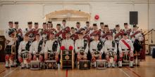 A pipe band poses for a group photo in a gym, showcasing their uniforms and instruments with smiles and camaraderie.
