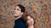The image is a medium shot portrait of a man and a woman positioned back-to-back against a colorful, mosaic tiled wall. The man is on the left, facing upwards and slightly to the left, with his head tilted back. He has short, dark brown hair, fair skin, and striking blue eyes. He is wearing a dark navy blue collared shirt.