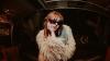 The image is a medium shot of a young woman posing in a dimly lit, somewhat cluttered indoor space. The woman is in the center of the frame, looking directly at the camera. She has reddish-brown hair with bangs and is wearing large, dark sunglasses with a thick black frame. Her left hand is resting on her cheek, with her fingers gently touching her face. She is wearing a light-colored, fluffy, faux fur jacket that covers most of her upper body. Several rings are visible on her left hand, and a beaded bracel