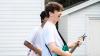 The image captures two young men outdoors, positioned in front of a white clapboard-sided building, likely a residential structure. The composition is dynamic, with the primary subject—a young man with tousled brown hair—occupying the right side of the frame. 