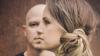 The image is a close-up, profile shot of two people, a man and a woman, superimposed or layered in a way that suggests a connection or duality. The man is bald and in the background, seen from the nose up, facing left. His expression is neutral. The woman is in the foreground, facing left, with her head angled slightly downwards. Her hair is light brown with blonde highlights, pulled back into a messy bun at the nape of her neck. Her makeup is visible, with eyeliner and mascara accentuating her eyes. 