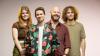 A group of four young adults, two women and two men, stand in a line facing forward against a neutral, light grey to pink gradient background. The lighting is warm and falls primarily on the faces and upper bodies of the subjects, creating soft shadows.