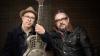 The image is a medium shot portrait of two middle-aged men, positioned side-by-side and looking towards the camera. They are both wearing glasses and appear to be musicians, as one of them is holding a resonator guitar.