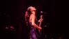 The image captures a dynamic moment of a female musician performing on stage. She is positioned in the center-left of the frame, looking upwards with her head tilted back, her eyes closed, exuding a sense of passionate immersion in her performance. Her dark, wavy hair is tied back into a ponytail and flows loosely around her shoulders and back.