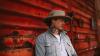 The image is a medium shot portrait of a man leaning against a weathered, corrugated red surface. The man is wearing a light grey, possibly corduroy, jacket over a white t-shirt. He has on a wide-brimmed, light brown hat. A prominent tattoo of a Native American headdress and profile is visible on his neck, extending upwards towards his ear. He is also wearing a necklace with multiple dark beads and a turquoise pendant.