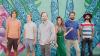 The image shows a group of six people standing side-by-side in front of a vibrant, colorful mural. The mural features intricate blue and green patterns that resemble mandala or floral designs, adding a striking artistic backdrop.