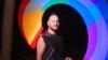 The image is a medium close-up portrait of a man standing in front of a blurred, circular light display at night. The man is looking directly at the camera with a slight smile. He has short, spiky dark hair, a beard, and is wearing a sleeveless, black sequined top and a silver chain necklace. His left arm is slightly extended, and he appears to be holding something small and dark in his hand.