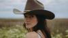 The image is a close-up portrait of a young woman wearing a wide-brimmed, dark brown cowboy hat. She is looking over her left shoulder towards the camera, with her face angled slightly to the right. Her expression is calm and direct, with a hint of a gaze.