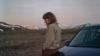 A young woman stands in profile beside the front of a dark-colored car, positioned slightly to the right of the frame. She has shoulder-length, wavy brown hair with bangs, and her head is tilted downward, her gaze directed toward the ground, suggesting introspection or melancholy.