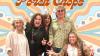 The image is a vibrant, medium shot of five people dressed in 1970s-inspired fashion, posing in front of a psychedelic, colorful backdrop. The backdrop features large, wavy bands of color in pastel shades of blue, light orange, and cream, interspersed with circles and starburst shapes, reminiscent of the era's art and design.