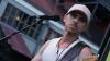 A male musician is performing on stage, holding an electric guitar and singing into a microphone. He is wearing a white tank top, a light pink baseball cap, and two silver necklaces with pendants. The guitar is a light brown color with a dark fretboard. The microphone is positioned in front of his face. The background is blurred but appears to be an outdoor venue with some structures and lights. The overall lighting suggests it is either daytime or early evening. The composition is a medium shot focusing on