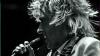 This is a black and white, close-up, profile shot of a male singer performing on stage. He has a full head of wild, spiky, blonde hair that frames his face and extends outward, suggesting movement. His head is tilted slightly downwards and to the left as he sings into a microphone held in his right hand. The microphone is dark and has a ribbed texture on its head. His mouth is open, and his eyes are not visible due to the angle.