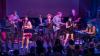 This is a medium shot of a live band performing on stage, captured under dramatic purple and blue stage lighting. The band consists of at least eight members, all actively engaged in their musical performance.
