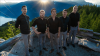 The image captures a group of five men standing on a rocky, gravelly overlook, seemingly in a mountainous region. The sun is setting or rising, casting a strong golden light with prominent sunbeams radiating from the top center of the frame, illuminating the scene
